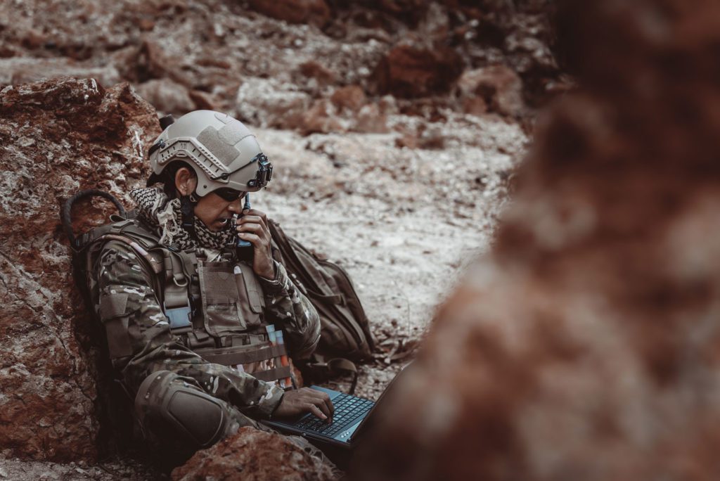 Soldier sitting on rocky surface using laptop and radio